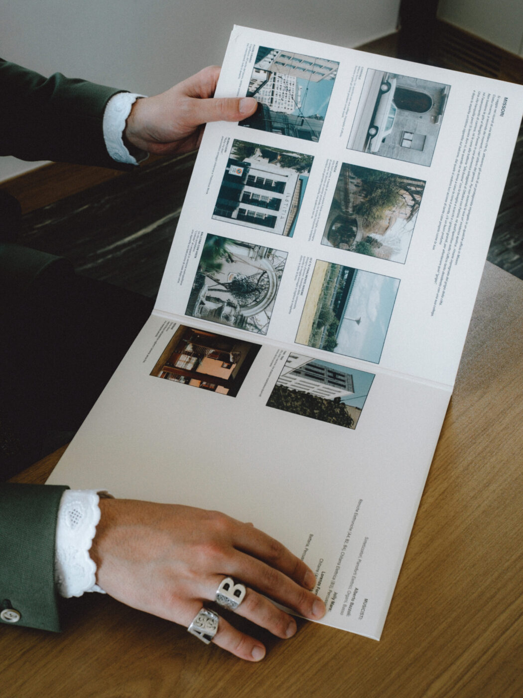A person wearing a green jacket and silver rings holds open a magazine or book featuring several photographs of buildings and outdoor scenes on a wooden table. *If you provide an image title, I’ll include it at the beginning as requested. Since none is present here, the alt text begins with the description only.*.