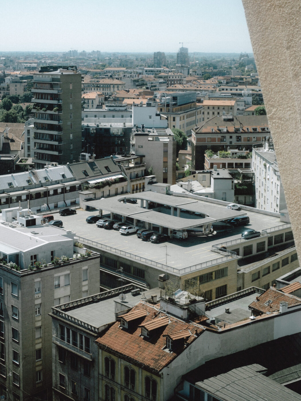 A cityscape view features mid-rise buildings with red-tiled roofs and a large rooftop parking lot with parked cars, set against a hazy, distant skyline under a clear sky.