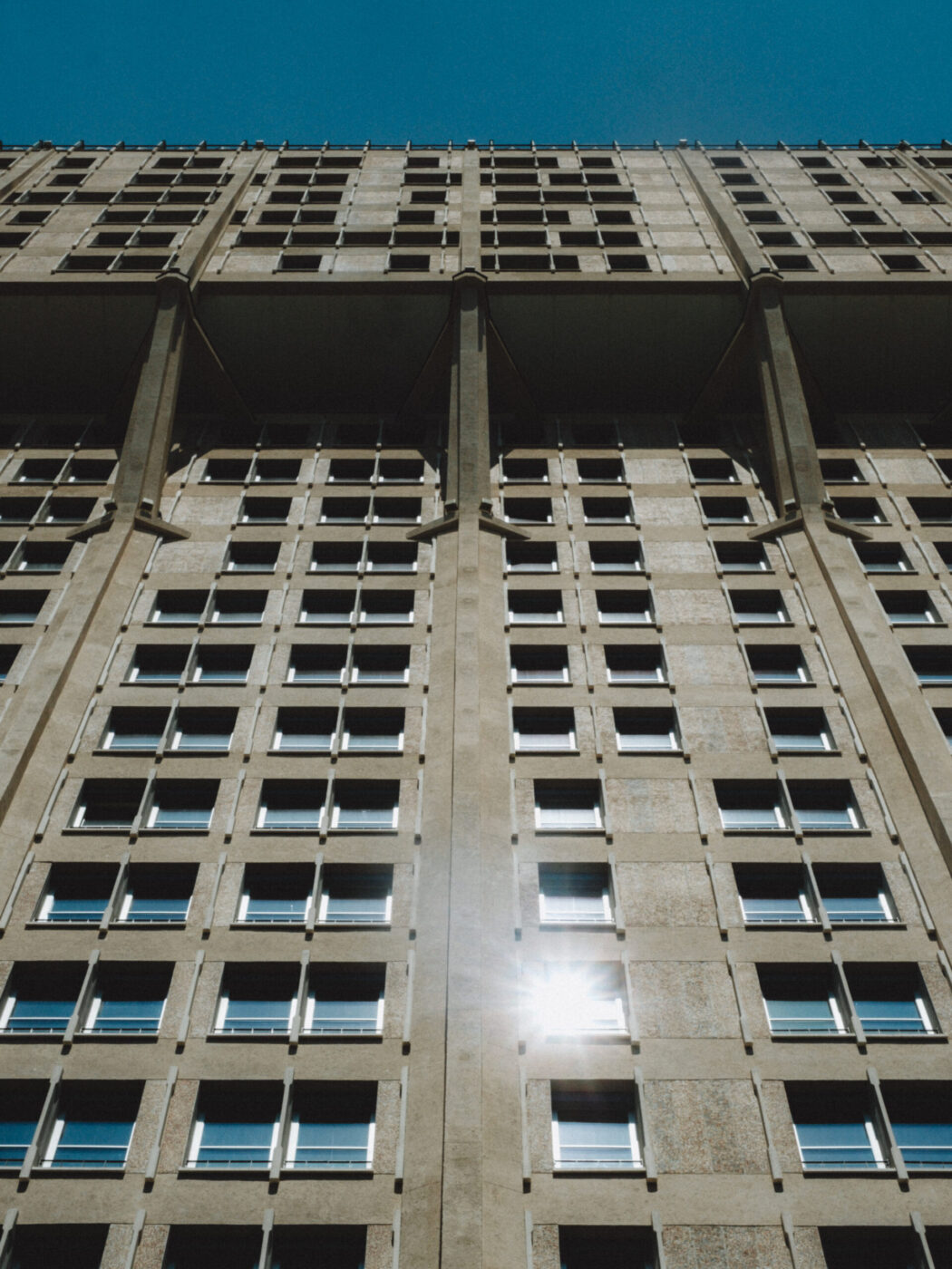 A tall concrete building with many rectangular windows, viewed from below against a clear blue sky. Sunlight reflects off one of the windows near the bottom center of the image.