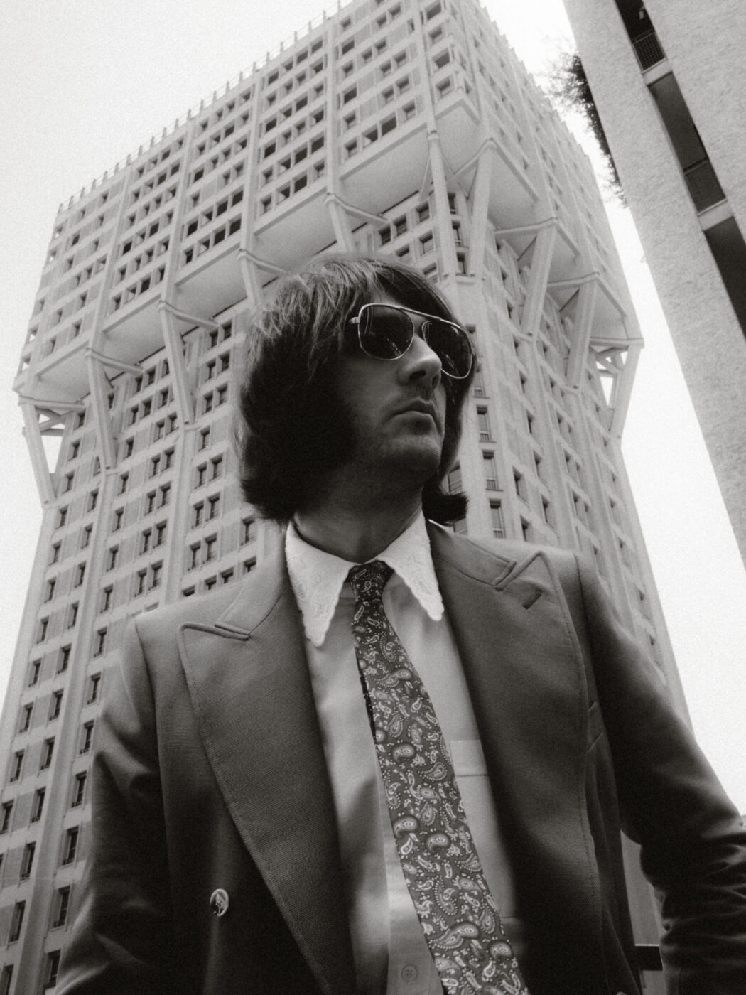 A man in a suit, patterned tie, and sunglasses stands in front of a tall, modern building with geometric architecture. The photo is black and white and taken from a low angle, emphasizing the building’s height.