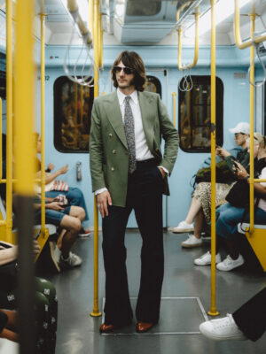 A man in sunglasses, a green blazer, and flared black pants stands confidently in the aisle of a subway car, surrounded by seated passengers.