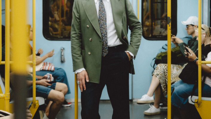Un uomo con occhiali da sole, blazer verde, camicia bianca e cravatta a fantasia sta in piedi con sicurezza nel corridoio di un treno della metropolitana, circondato da passeggeri seduti.