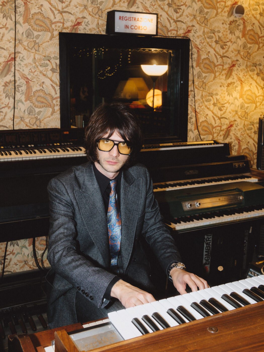 A person in a gray suit and patterned tie plays a vintage keyboard in a retro-styled room with patterned wallpaper, multiple keyboards in the background, and a sign above reading REGISTRAZIONE IN CORSO.
