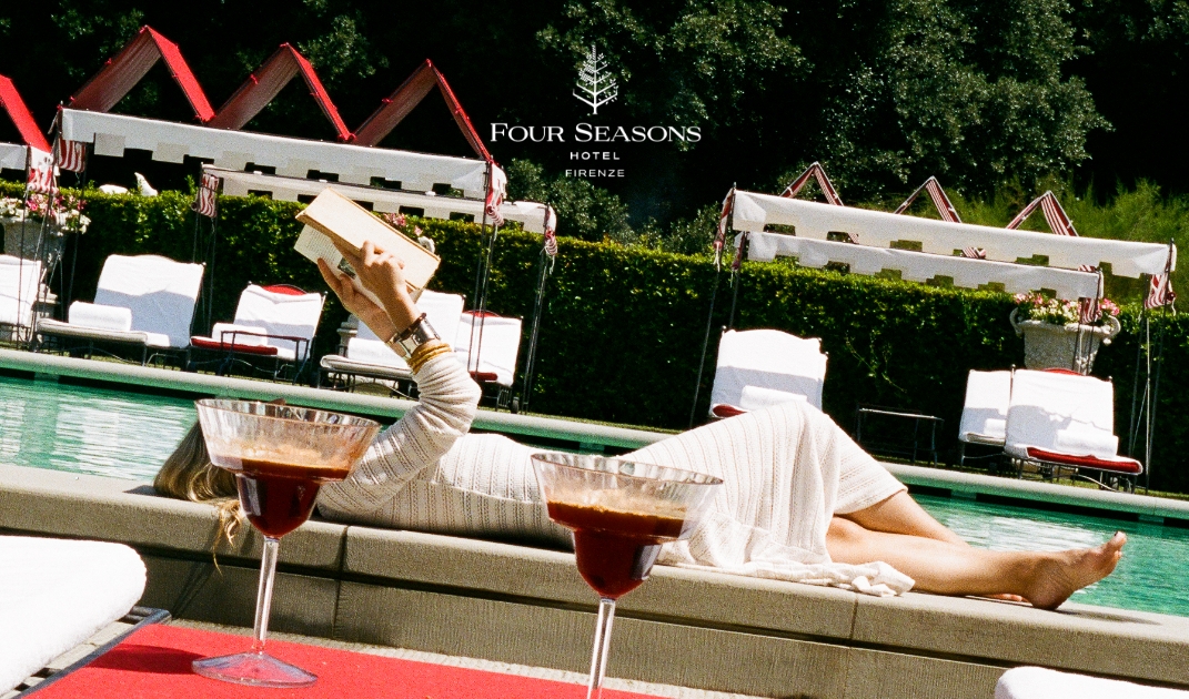 Certainly! Here’s the image alt text following your instructions: A person in a white dress lounges by a pool at the Four Seasons Hotel Firenze - A person in a white dress lounges by a pool at the Four Seasons Hotel Firenze, holding a book overhead. Two cocktails sit in the foreground on a red mat, with cabanas and greenery in the background.