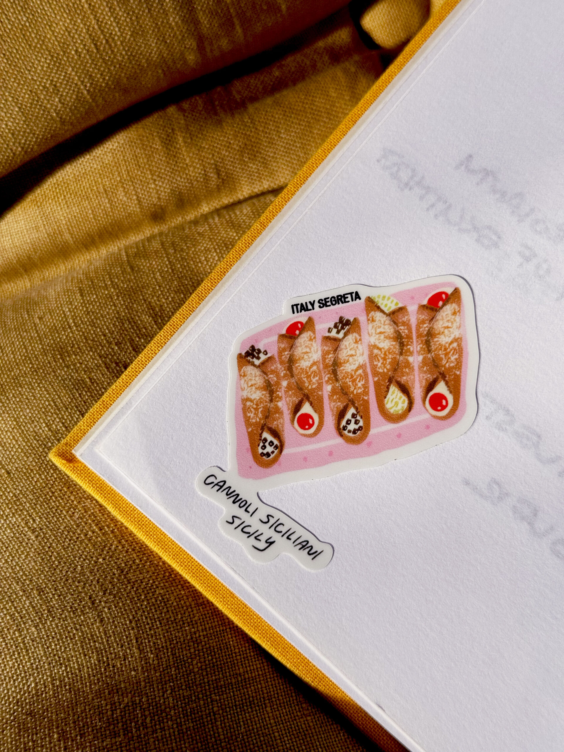 Cannoli Siciliani Sicily - An A Tavola Stickers sheet featuring five cannoli with varied toppings sits inside a card labeled CANNOLI SICILIANI SICILY, all set against a gold fabric background.