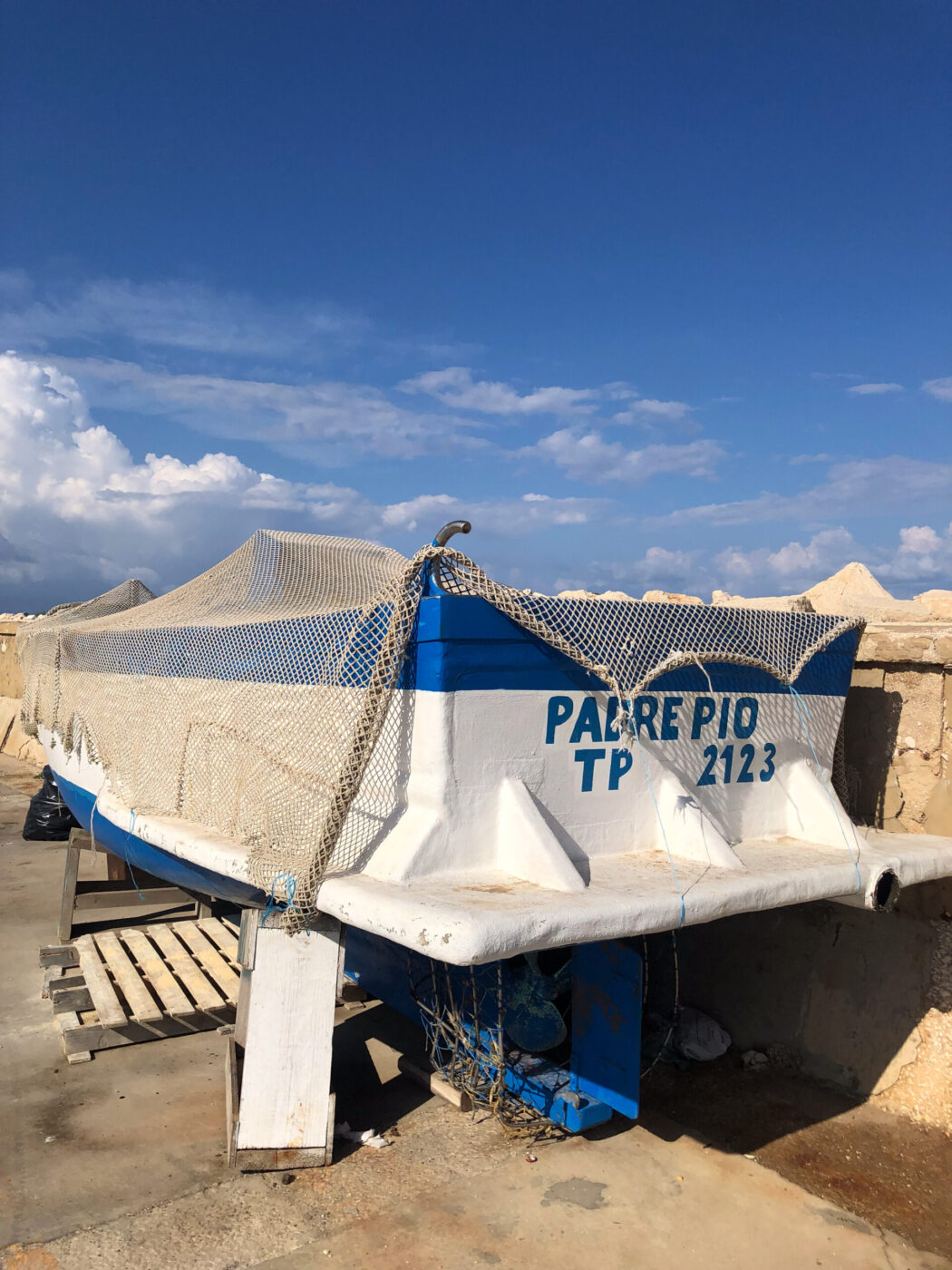 PADRE PIO TP 2123 - Un peschereccio bianco e blu di nome PADRE PIO TP 2123 si trova sulla terraferma accanto a un muro di pietra, coperto da una rete sotto un cielo azzurro e limpido con nuvole sparse.