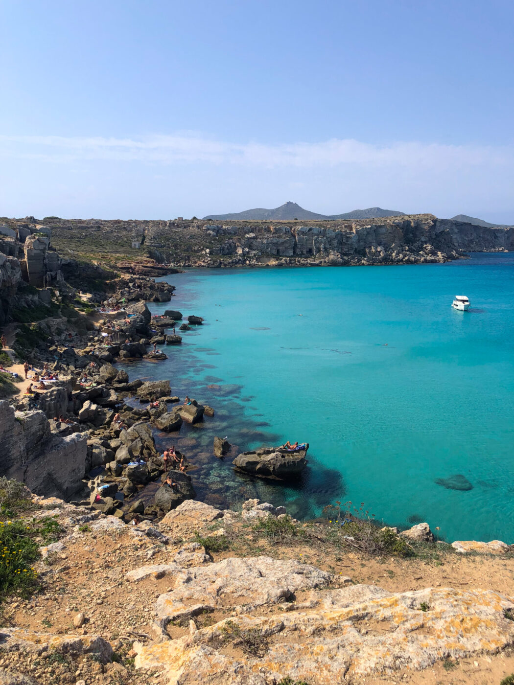 Una costa rocciosa con acqua limpida e turchese, persone che prendono il sole sugli scogli e una barca bianca ancorata nella baia; colline lontane sotto un cielo azzurro.