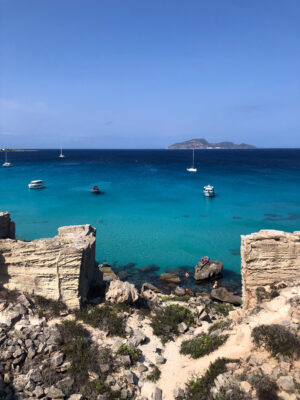 View of a turquoise sea with boats anchored, rocky ruins and shrubs in the foreground, and a distant island under a clear blue sky. Swimmers can be seen in the shallow water near the shore. If no image title is available (as in your prompt), you would just use: View of a turquoise sea with boats anchored, rocky ruins and shrubs in the foreground, and a distant island under a clear blue sky. Swimmers can be seen in the shallow water near the shore. If youd like me to generate an example with another specific title or need further help formatting more examples this way, just let me know!.