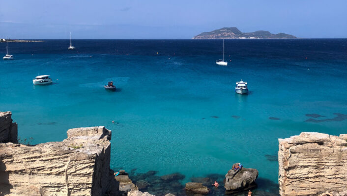 View of a turquoise sea with boats anchored, rocky ruins and shrubs in the foreground, and a distant island under a clear blue sky. Swimmers can be seen in the shallow water near the shore. If no image title is available (as in your prompt), you would just use: View of a turquoise sea with boats anchored, rocky ruins and shrubs in the foreground, and a distant island under a clear blue sky. Swimmers can be seen in the shallow water near the shore. If youd like me to generate an example with another specific title or need further help formatting more examples this way, just let me know!.
