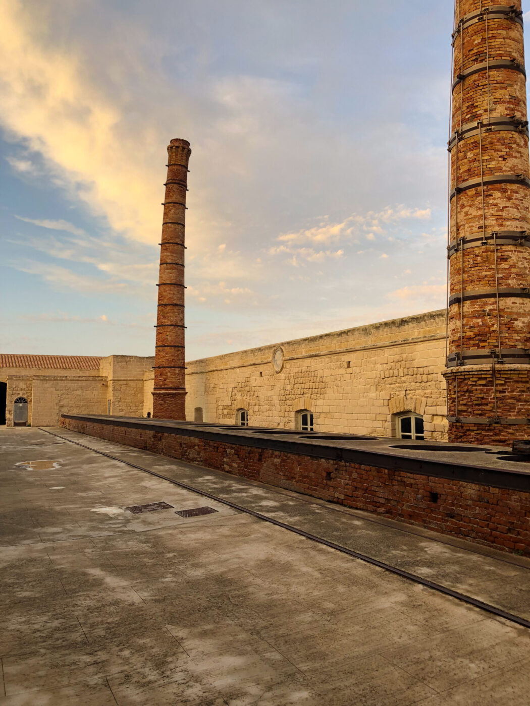 [Titolo immagine] - Due alte ciminiere cilindriche in mattoni si ergono da un edificio industriale in pietra sotto un cielo di nuvole soffici, proiettando lunghe ombre su un cortile di cemento vuoto.