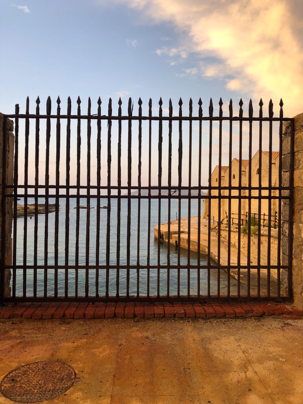 Non è stato fornito un titolo per l'immagine. Testo Alt: Un'alta recinzione di ferro blocca l'accesso a un molo di pietra che si estende in un mare azzurro e calmo, con edifici lungo il molo e un cielo parzialmente nuvoloso durante il tramonto.