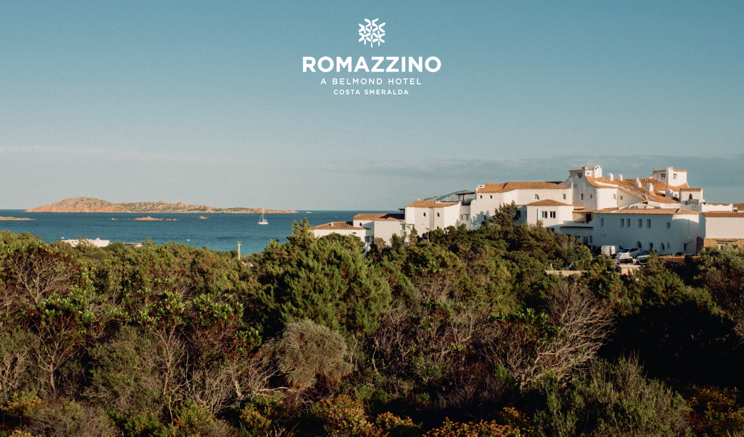 Romazzino, un hotel Belmond in Costa Smeralda - Una vista costiera di Romazzino, un hotel Belmond in Costa Smeralda, con edifici bianchi e tetti in terracotta che si affacciano sul mare blu, circondati da una vegetazione lussureggiante sotto un cielo limpido.
