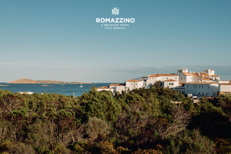 Romazzino, A Belmond Hotel, Costa Smeralda - Vista di edifici bianchi con tetti in terracotta vicino al mare, circondati da una vegetazione verde sotto un cielo azzurro e limpido. L'immagine include il logo Romazzino, A Belmond Hotel, Costa Smeralda in alto.