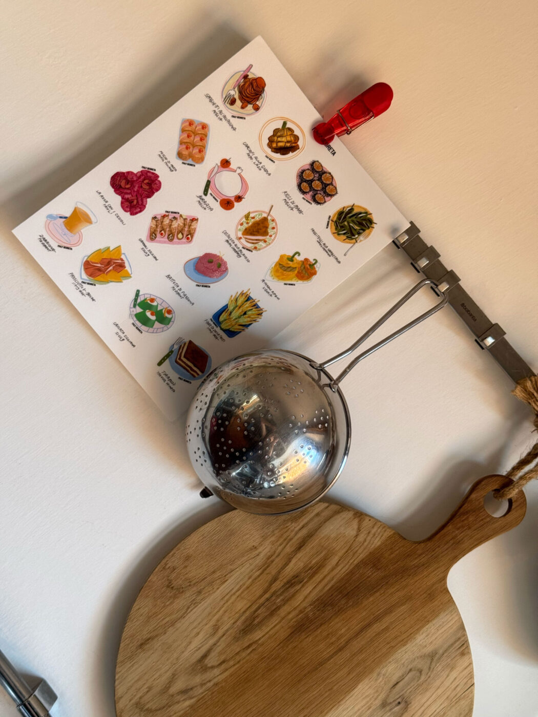 A Tavola Stickers - Colorful illustrated kitchen utensils stickers are displayed on a light wall, positioned above a metal strainer and a round wooden cutting board with a handle.