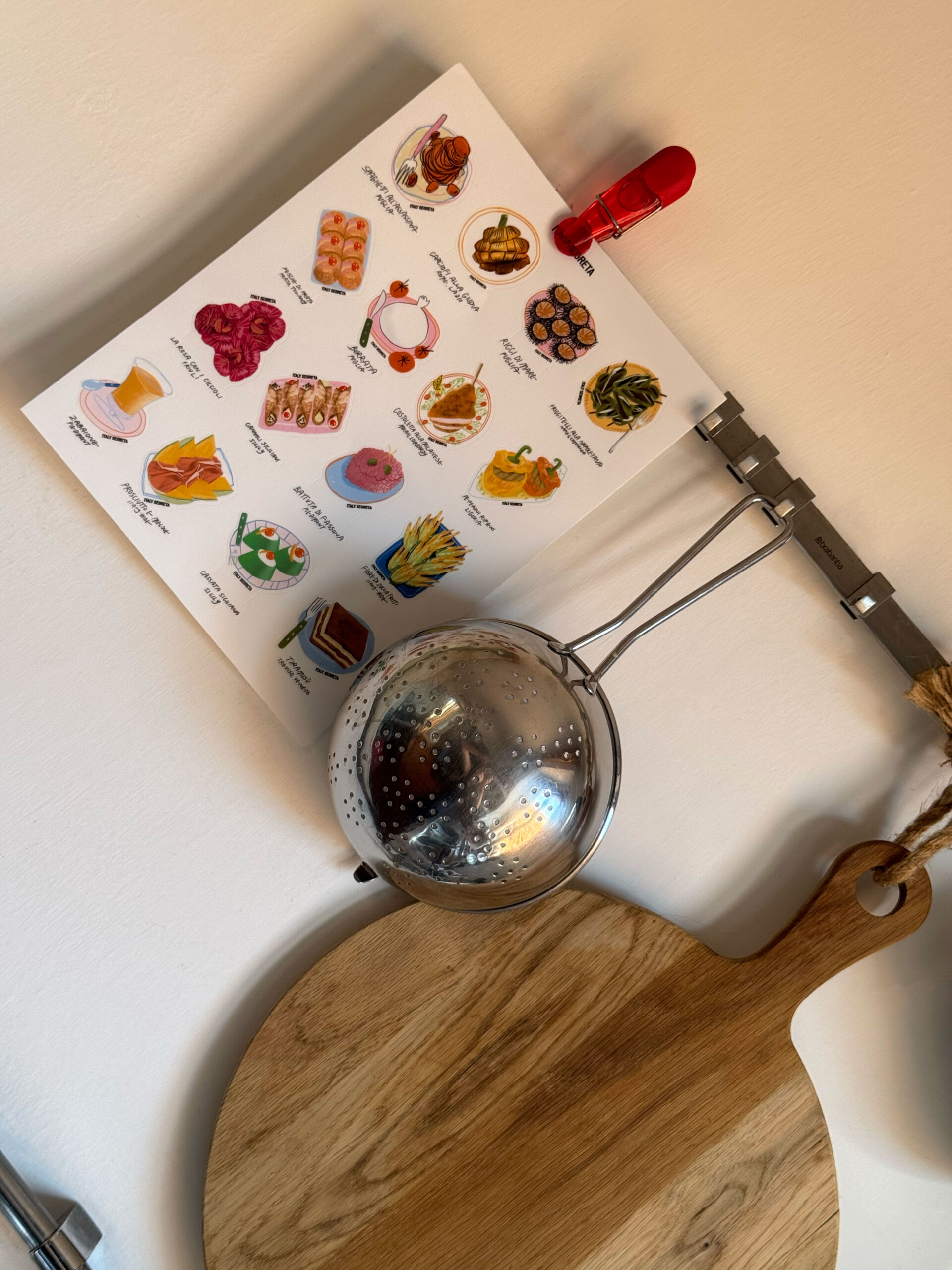 A Tavola Stickers - Colorful illustrated kitchen utensils stickers are displayed on a light wall, positioned above a metal strainer and a round wooden cutting board with a handle.