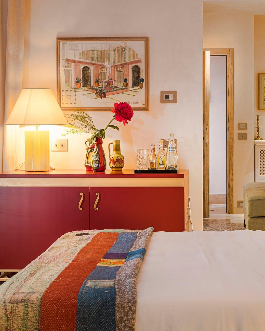 There is no image title provided. Alt text: A cozy bedroom features a bed with a colorful quilt, a red cabinet topped with a lamp, vases, and drinks, and a painting on the wall. A doorway leads to another room, and warm lighting creates an inviting atmosphere.