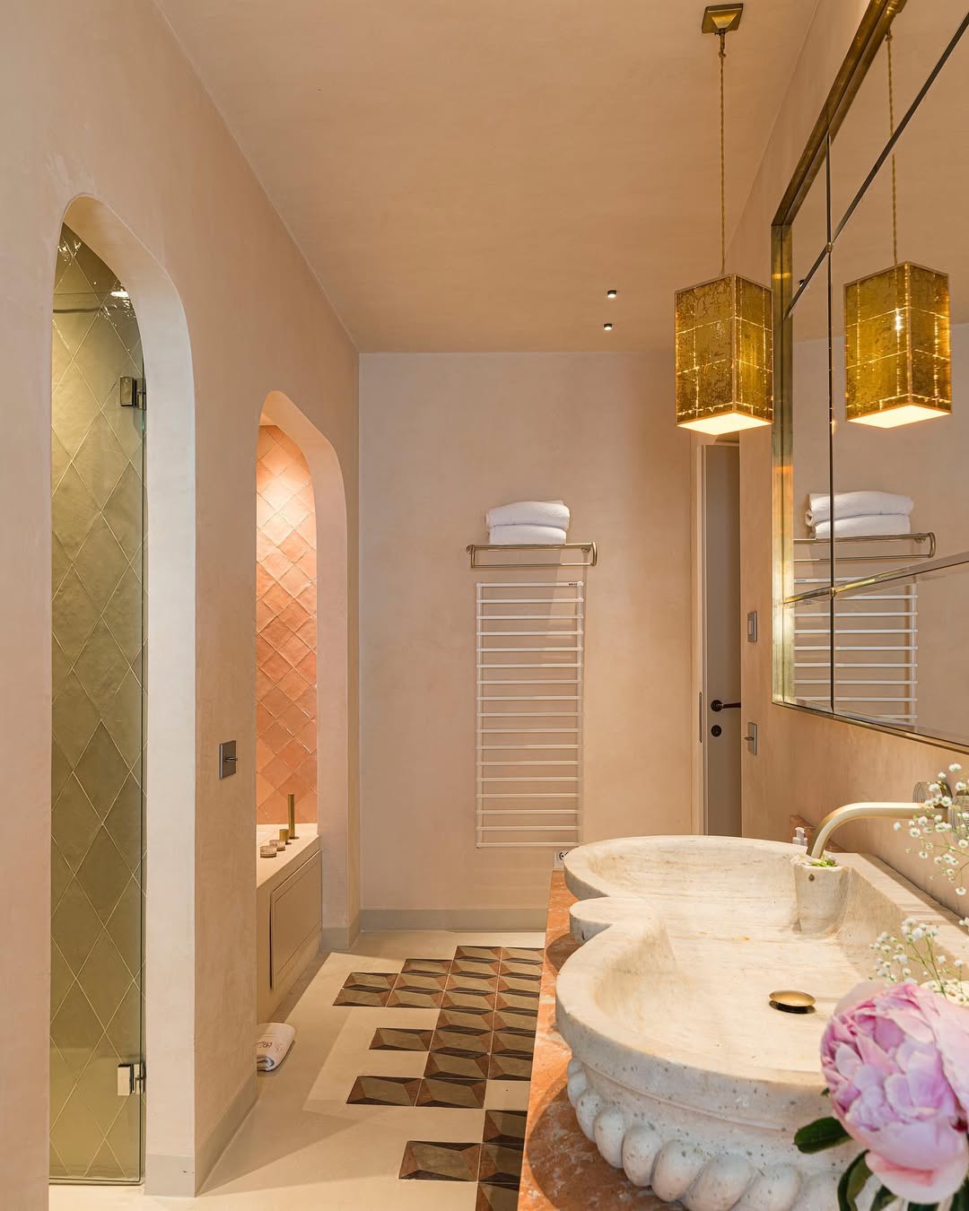 Elegant bathroom with a marble double sink, golden hanging lights, large mirror, towel rack, glass shower, bathtub with pink tiles, and decorative floor tiles - An elegant bathroom featuring a wide marble double sink counter topped with a vase of flowers. Golden hanging pendant lights illuminate the area above the large wall mirror. A towel rack is nearby. To one side sits a glass-enclosed shower; adjacent is a bathtub accented by soft pink tiles. The floor displays intricate decorative tiling throughout the space.