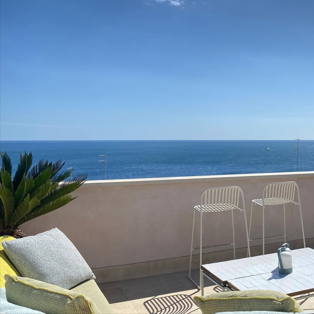 A sunny terrace with cushions, a small table, two white bar stools, a potted plant, and a clear view of the calm blue sea under a bright sky.
