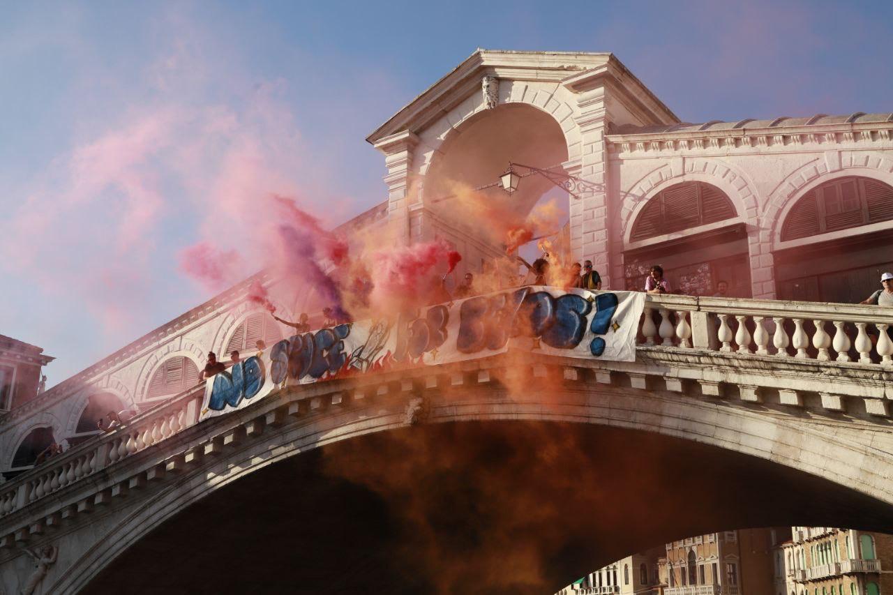 [Persone in piedi su un ponte di pietra che reggono uno striscione mentre intorno a loro si alzano nuvole di fumo arancione e rosa. Il ponte appare storico con l'architettura ad arco e la luce del sole che illumina la scena.