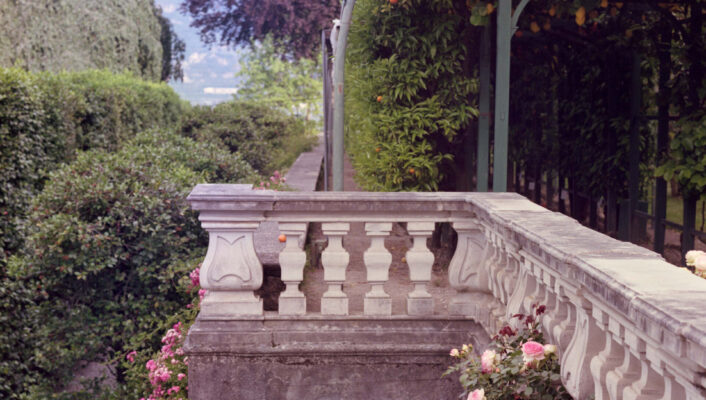 Terrazza in pietra con rose rosa sulla balaustra, siepi rigogliose, arco di vite a destra, montagne in lontananza sotto un cielo pallido.