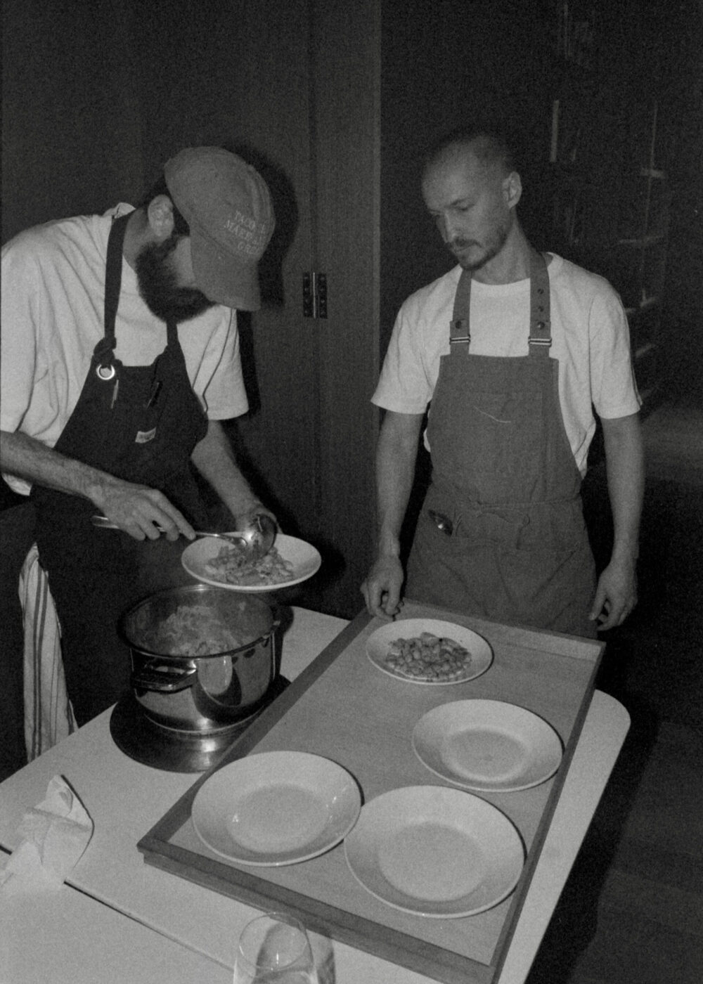 Based on your instruction, the alt text should be: If an image title is present, always add the image title at the beginning of the alt text. Format: [Image Title] - [Alt Text] : Two men in aprons prepare and serve food from a pot onto plates in a dimly lit kitchen; one scoops food while the other watches, and several empty plates are on a tray in front of them. Black and white image. If we assume Soup Kitchen Volunteers is an example image title, then according to your format: Soup Kitchen Volunteers - Two men in aprons prepare and serve food from a pot onto plates in a dimly lit kitchen; one scoops food while the other watches, and several empty plates are on a tray in front of them. Black and white image. Let me know if you need it adapted for another specific image title!.