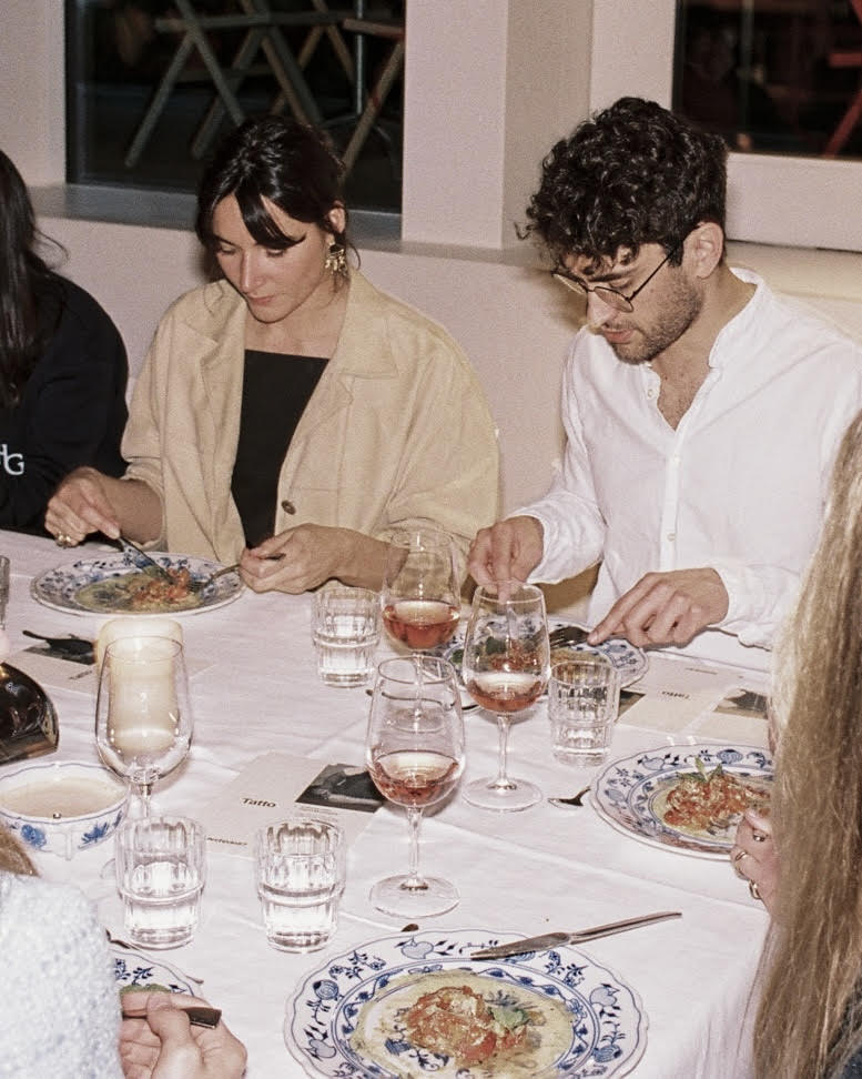 Table for Two - Two people sit at a table set with floral plates, wine glasses, and candles, enjoying a meal together. They appear focused on their food in a warmly lit, casual dining setting.