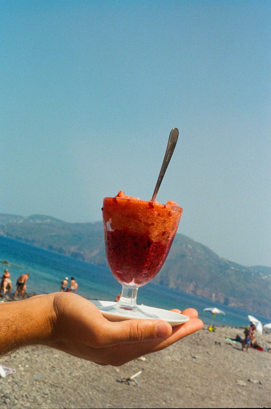 Certo! Ecco il testo alt nel formato richiesto: Una mano tiene un bicchiere di granita rossa e arancione - Una mano tiene un bicchiere di granita rossa e arancione con un cucchiaio, in una scena di spiaggia con persone, montagne e un cielo blu brillante sullo sfondo.
