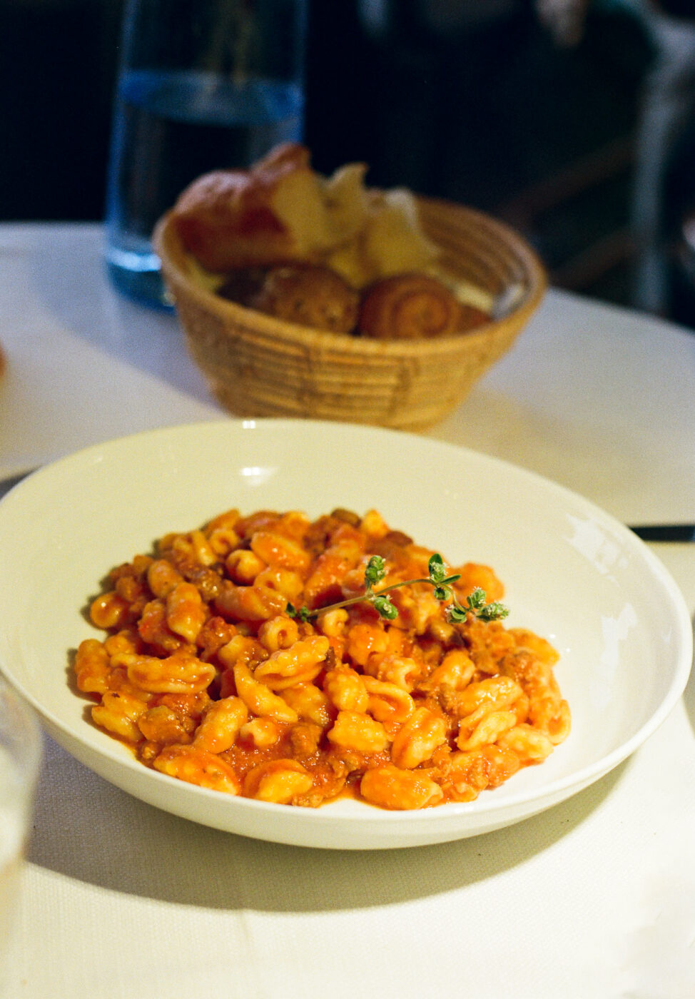 A white bowl with pasta in tomato sauce and thyme sits on a table; bread basket and water bottle are in the background.