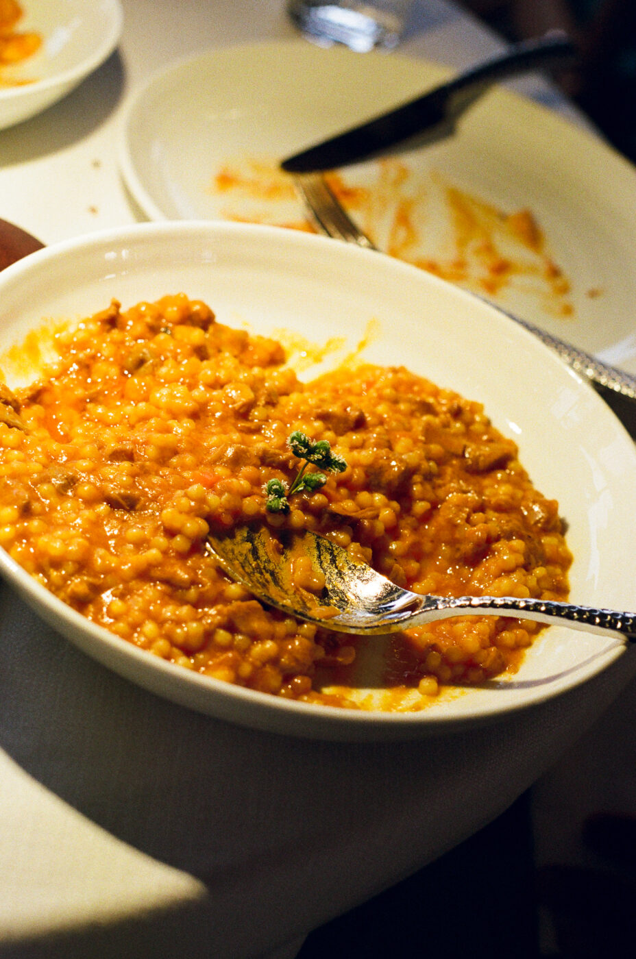 A bowl of orange fregola with a herb sprig, spoon in the dish, on a white tablecloth with an empty plate behind.