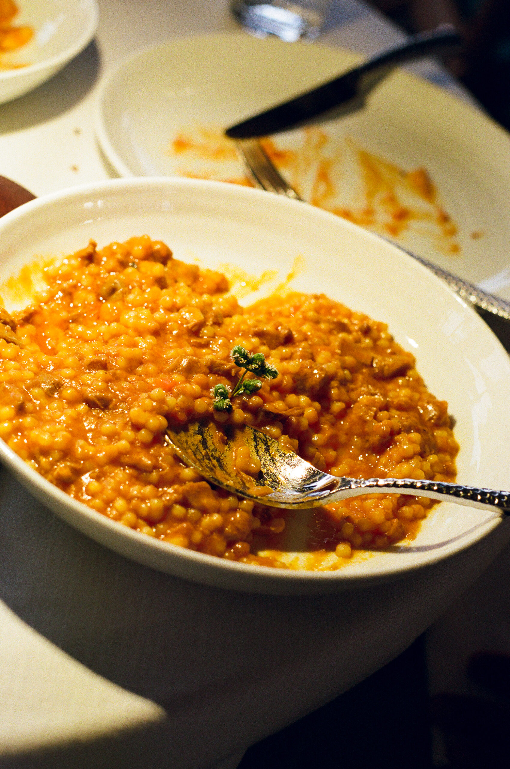 A bowl of orange fregola with a herb sprig, spoon in the dish, on a white tablecloth with an empty plate behind.