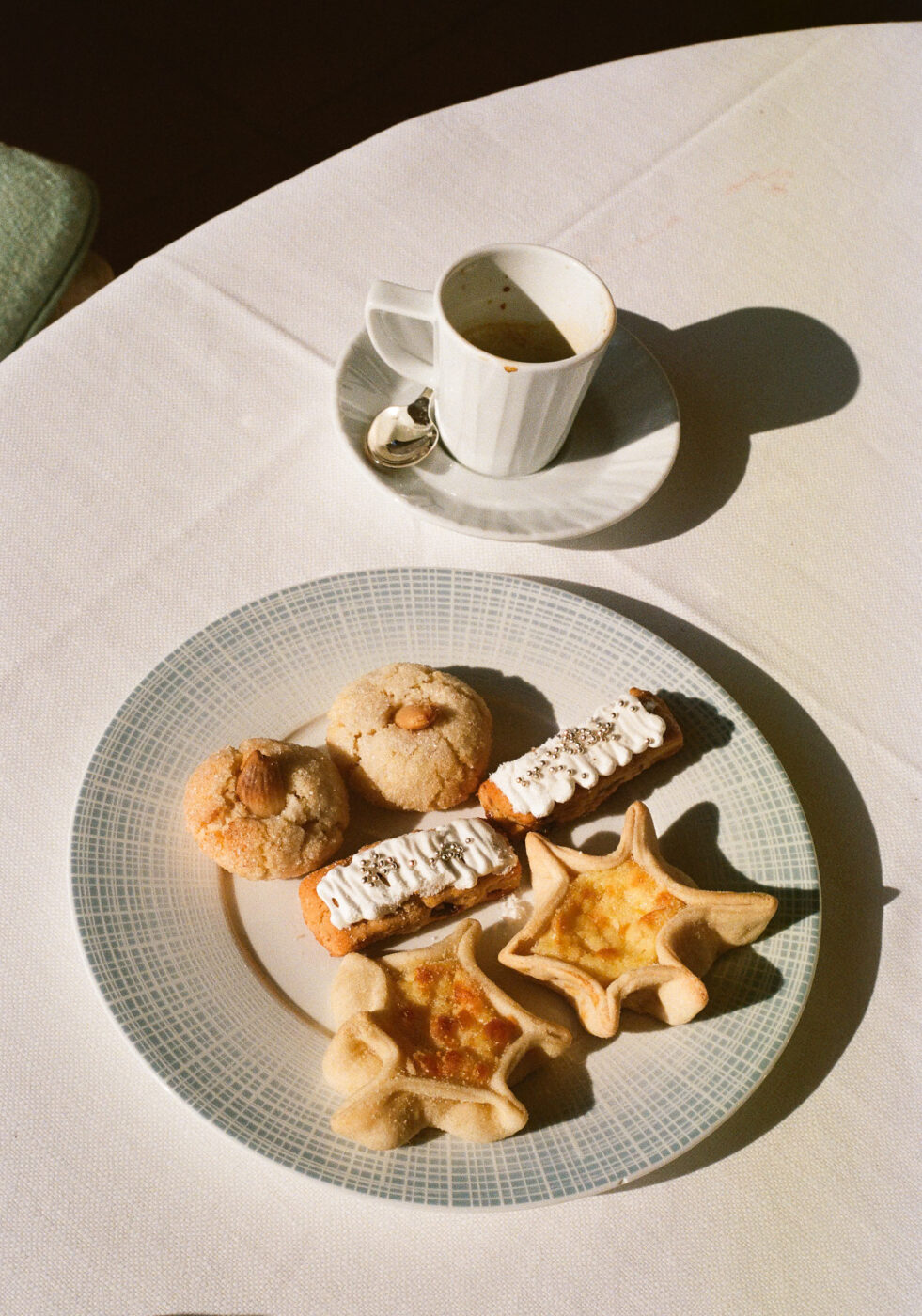 A white plate with assorted cookies and pastries on a white tablecloth, beside a white espresso cup; sunlight casts soft shadows.