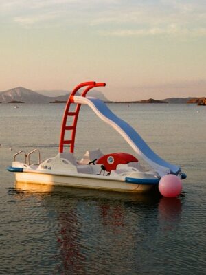 A small pedal boat with a red slide and a pink buoy – A small pedal boat with a red slide and a pink buoy floats on calm, clear water at sunset, with distant islands and a partly cloudy sky in the background.
