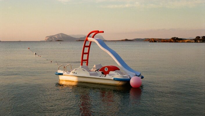 Non è presente alcun titolo dell'immagine, quindi il testo alt sarebbe: Un piccolo pedalò bianco con uno scivolo rosso e una boa rosa galleggia sull'acqua calma del mare al tramonto, con isole lontane e un cielo limpido sullo sfondo.