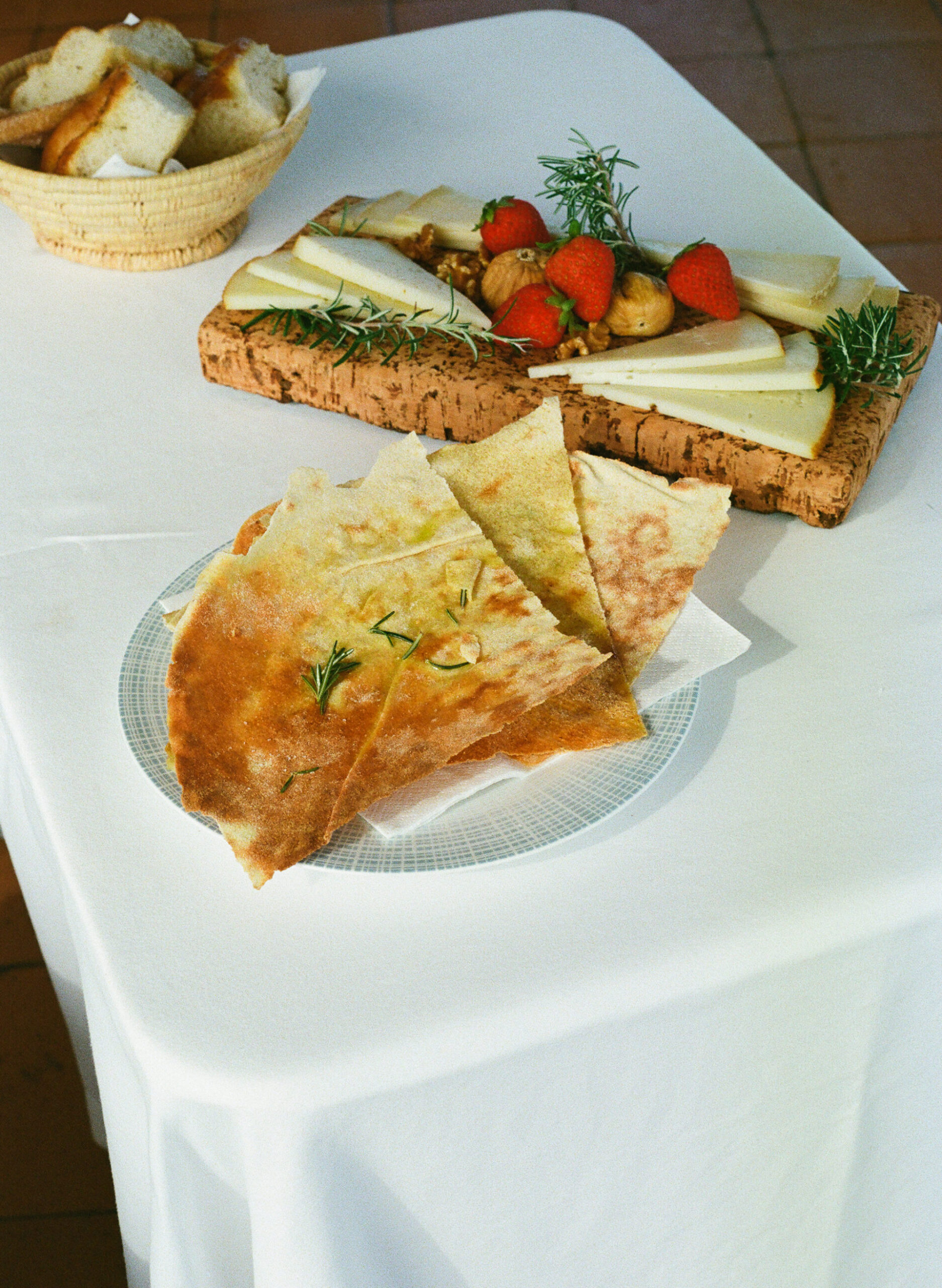 A plate of rosemary flatbread on a white tablecloth, with cheese, nuts, strawberries, and bread on wooden platters in back.