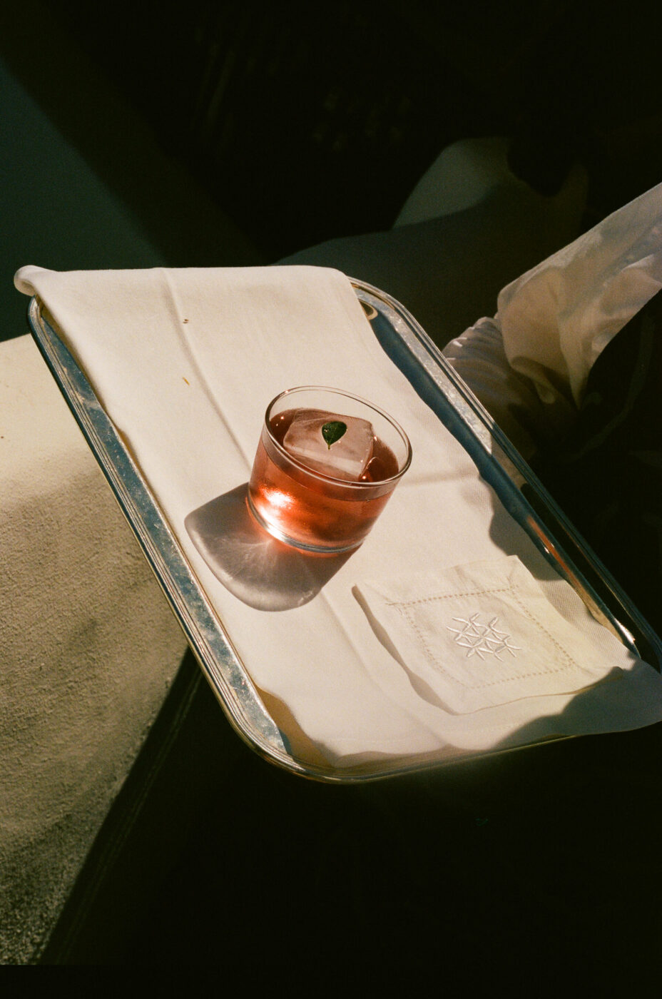 A glass of pink drink with a big ice cube and a leaf rests on a silver tray with a white napkin, glowing in the warm sunlight.