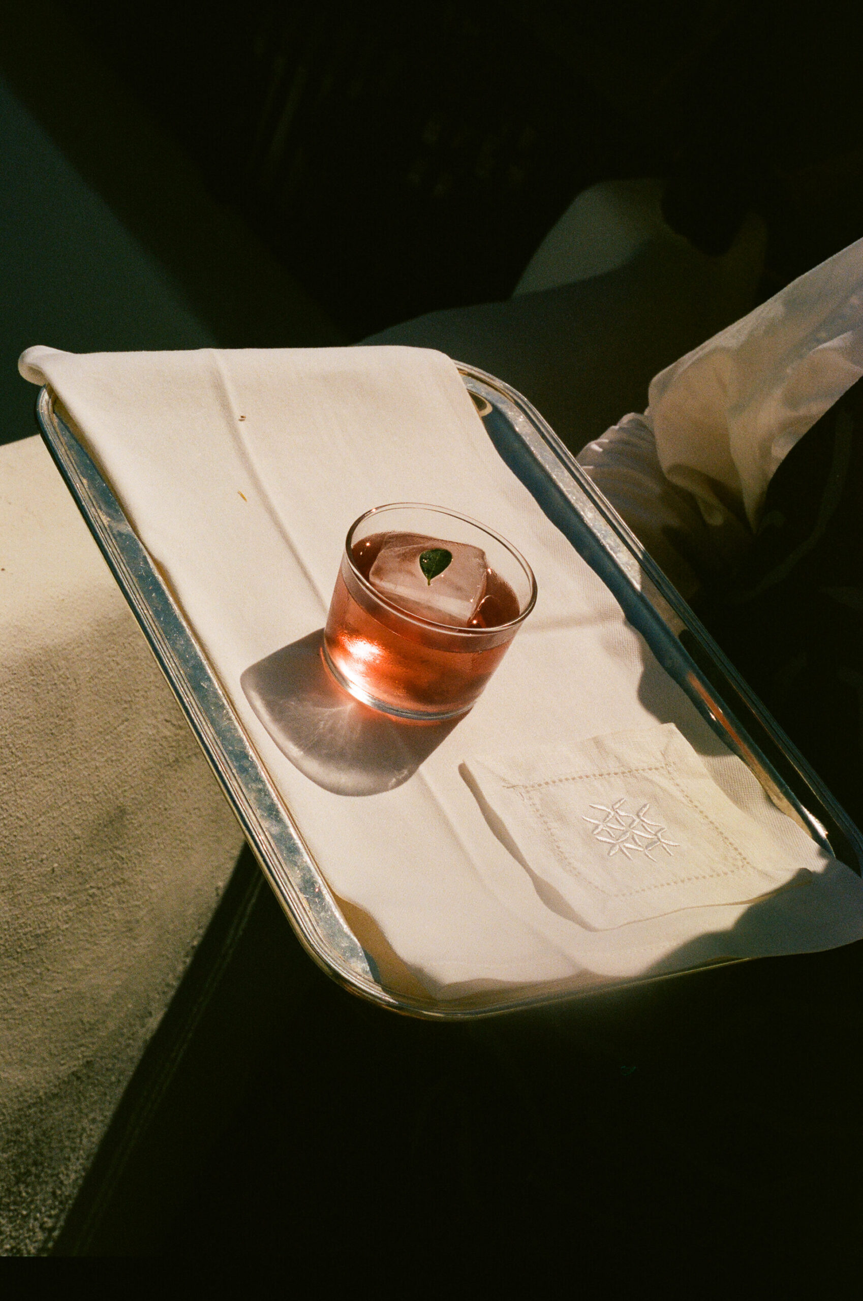 A glass of pink drink with a big ice cube and a leaf rests on a silver tray with a white napkin, glowing in the warm sunlight.