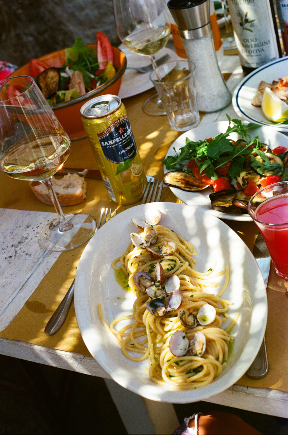 Pasto italiano all'aperto - Un tavolo apparecchiato all'aperto con piatti di spaghetti e vongole, un'insalata fresca, bruschette, due bicchieri da vino, una lattina di San Pellegrino, una fetta di pane e condimenti, il tutto immerso nella luce del sole.