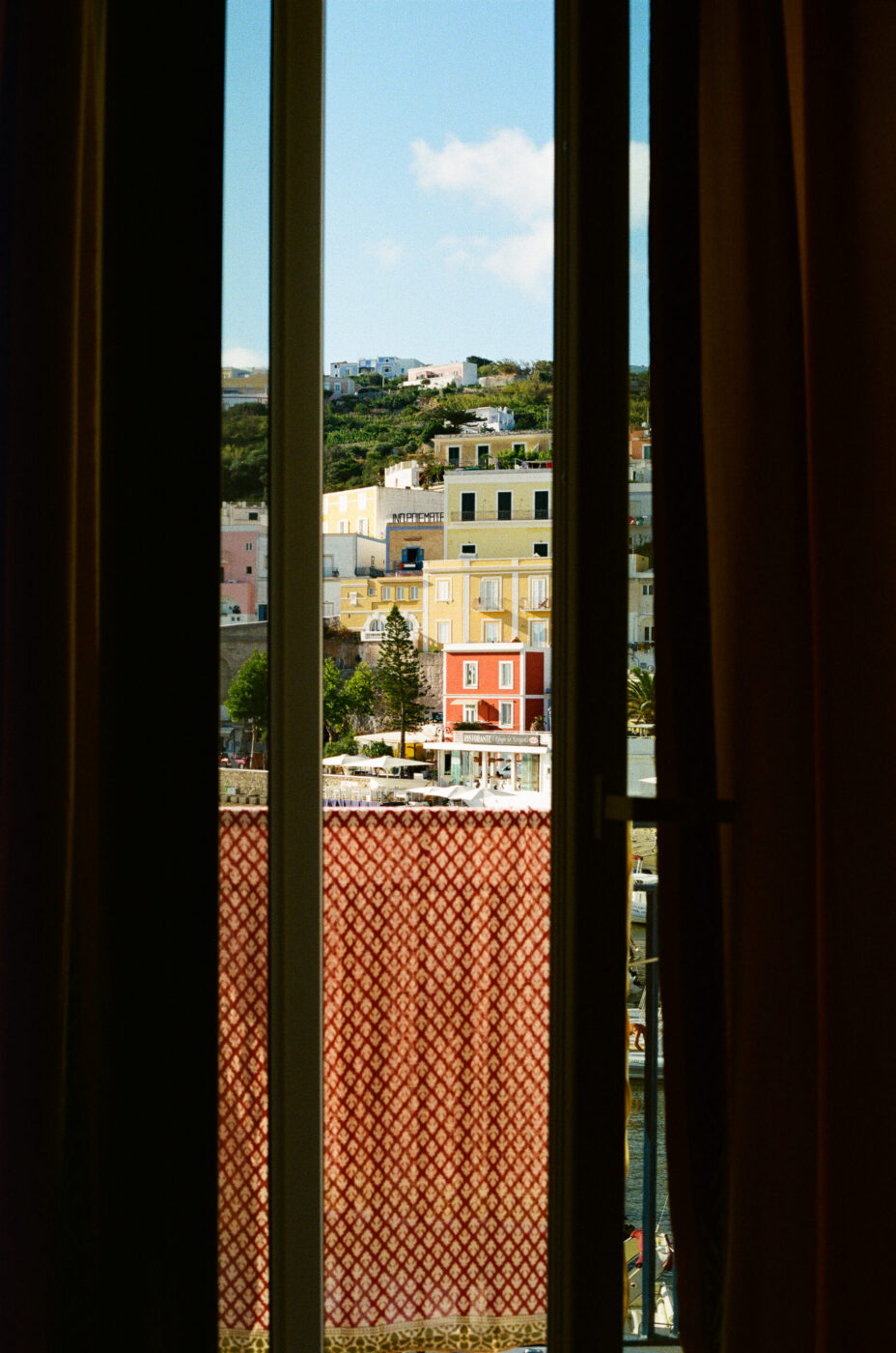 La vista attraverso una finestra incorniciata da tende scure mostra edifici collinari colorati e un balcone piastrellato, con un cielo azzurro e qualche nuvola sopra.