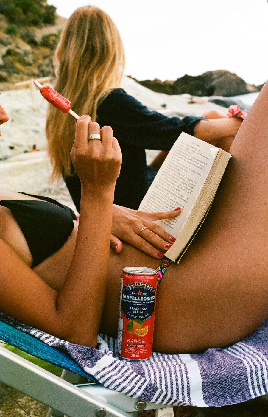 Una persona in bikini nero si sdraia su una sedia da spiaggia, leggendo un libro e tenendo in mano un ghiacciolo. Sulle sue ginocchia è appoggiata una lattina di Sanpellegrino; un'altra persona con lunghi capelli biondi siede vicino, rivolta verso l'esterno.