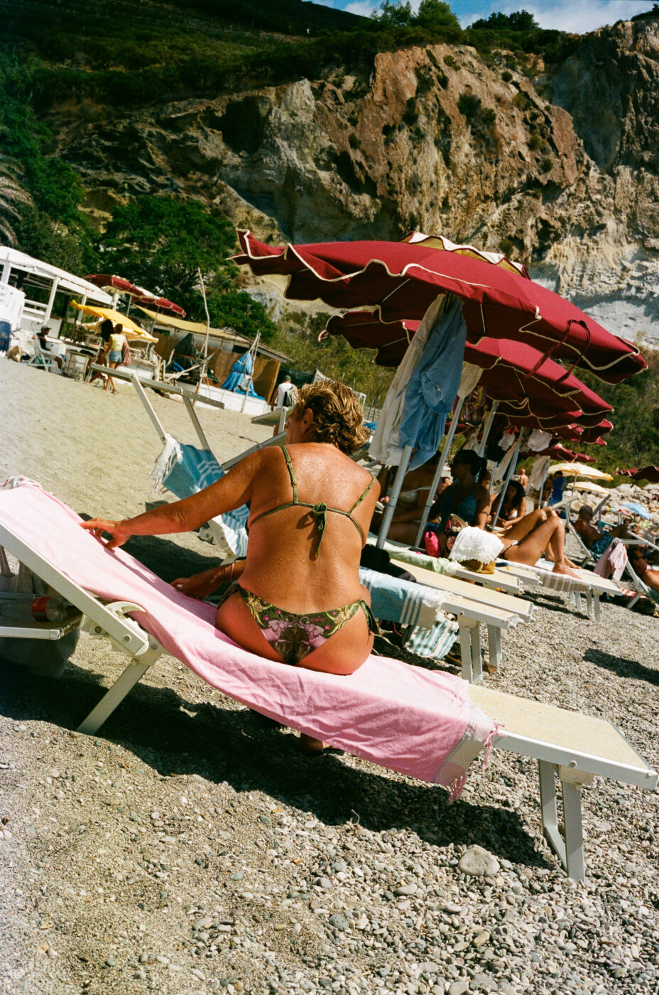 Sembra che si voglia generare il testo alt in base alle linee guida e alla descrizione fornite. Ecco il testo alt formattato: Una donna in bikini siede su una sedia a sdraio coperta da un asciugamano rosa in una spiaggia rocciosa. Ombrelloni rossi ombreggiano altri bagnanti nelle vicinanze. Sullo sfondo, sotto un cielo parzialmente nuvoloso, si ergono vegetazione e scogliere rocciose. Se il titolo dell'immagine è già presente, vi prego di fornirlo e io lo aggiungerò all'inizio secondo il vostro formato. Ad esempio, se il titolo dell'immagine è "Seaside Relaxation": Seaside Relaxation - Una donna in bikini siede su una sedia a sdraio coperta da un asciugamano rosa in una spiaggia rocciosa. Ombrelloni rossi fanno ombra ad altri bagnanti nelle vicinanze. Sullo sfondo, sotto un cielo parzialmente nuvoloso, si ergono vegetazione e scogliere rocciose.