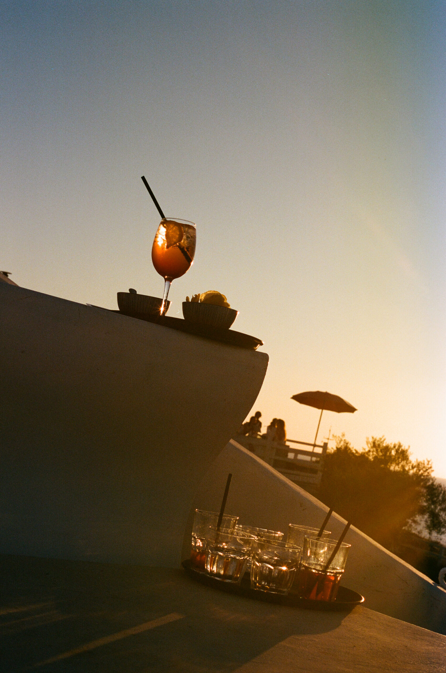 Sunset Cocktail - Un cocktail con una fetta di limone e una cannuccia si trova su un cornicione al tramonto, con diversi bicchieri vuoti e un'altra cannuccia su un vassoio sottostante. La scena è all'aperto con la luce calda del sole e le sagome sullo sfondo.
