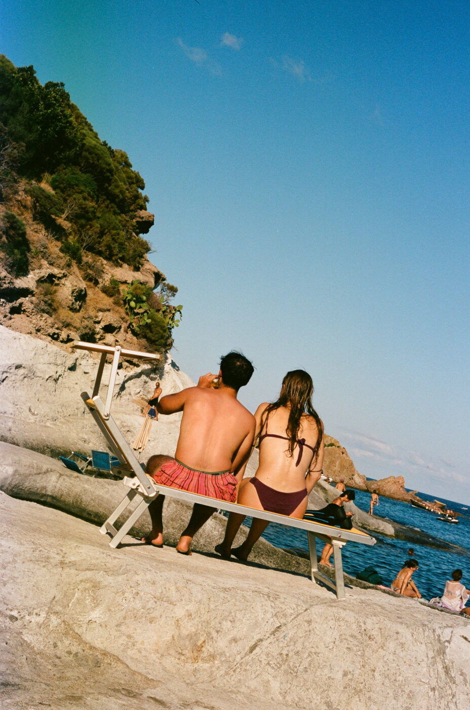 Certo! Ecco il testo alt seguendo le tue istruzioni: [Titolo immagine] - Due persone in costume da bagno siedono su una sedia a sdraio di fronte al mare, circondate da un terreno roccioso e da altri frequentatori della spiaggia. Il tempo è soleggiato con un cielo azzurro e limpido.