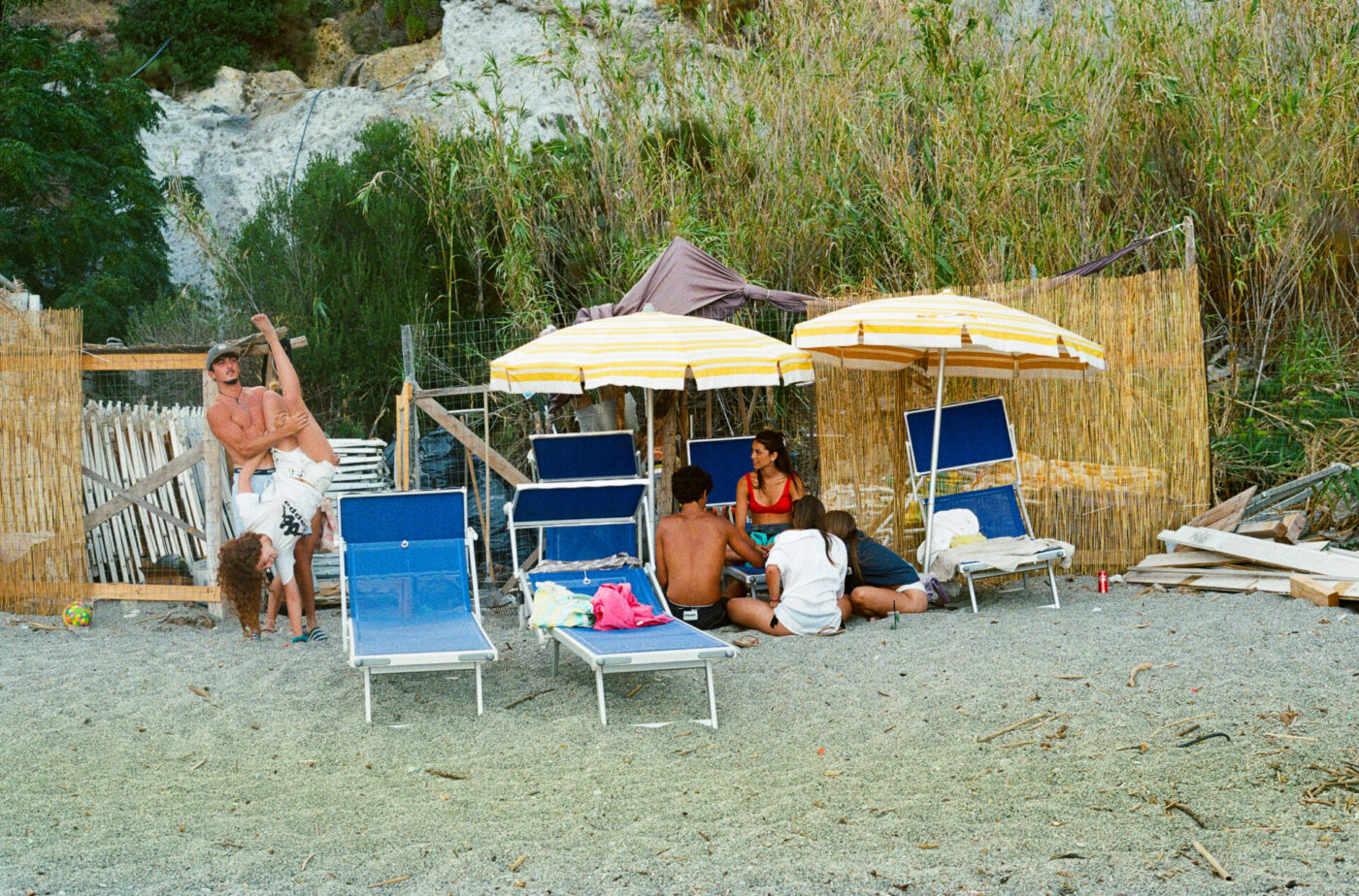 Un gruppo di persone si rilassa su una spiaggia rocciosa - Un gruppo di persone si rilassa su una spiaggia rocciosa con sedie a sdraio blu, ombrelloni a strisce gialle e paraventi di bambù. Due persone ballano giocosamente sulla sinistra, mentre altre quattro siedono e parlano vicino agli ombrelloni.