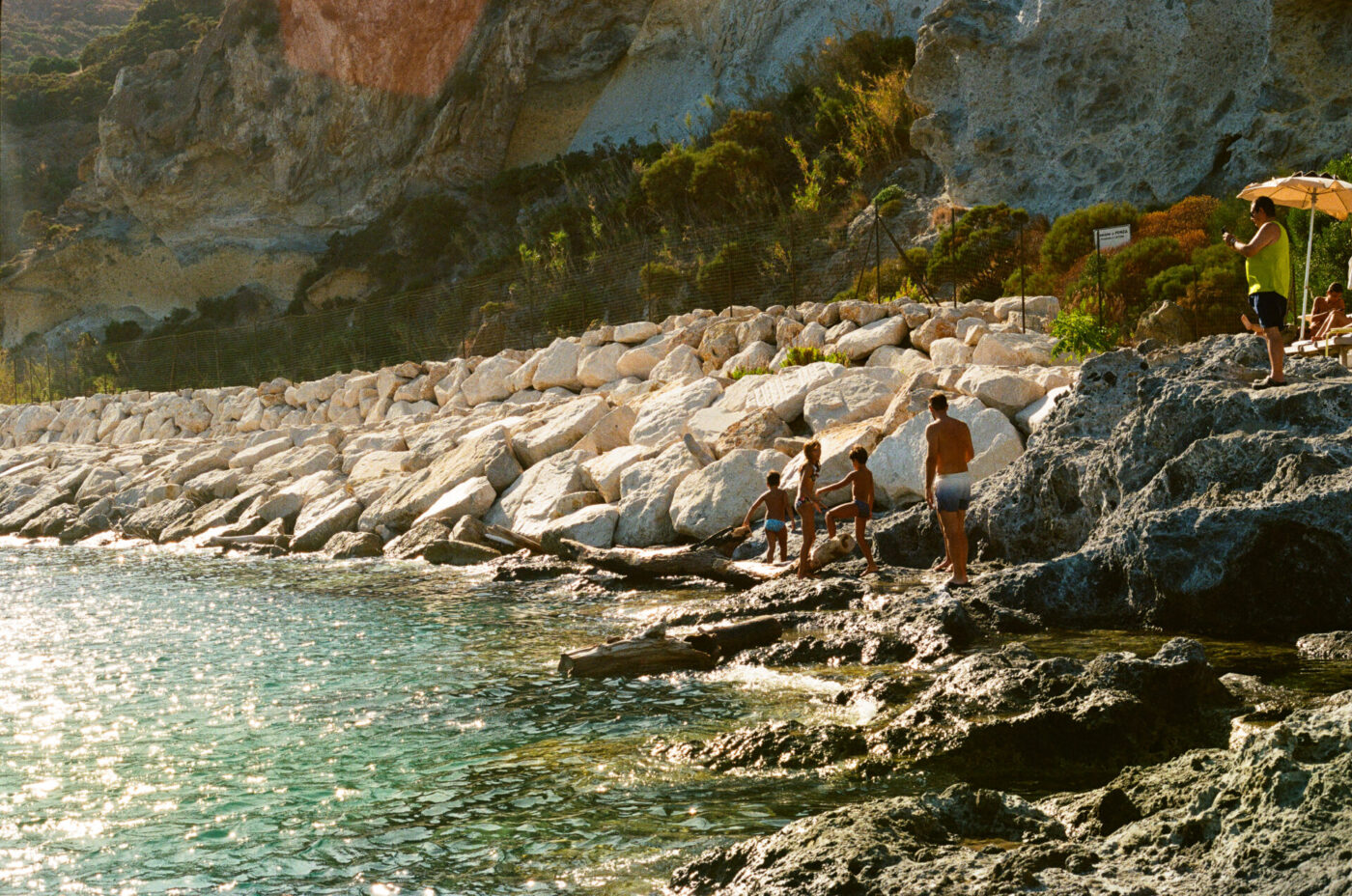 Titolo dell'immagine non fornito - Persone in piedi e sedute sulla riva rocciosa in riva all'acqua turchese, con la luce del sole che si riflette sul mare. Grandi pietre bianche formano una barriera lungo la costa e arbusti verdi crescono sulla scogliera sullo sfondo.