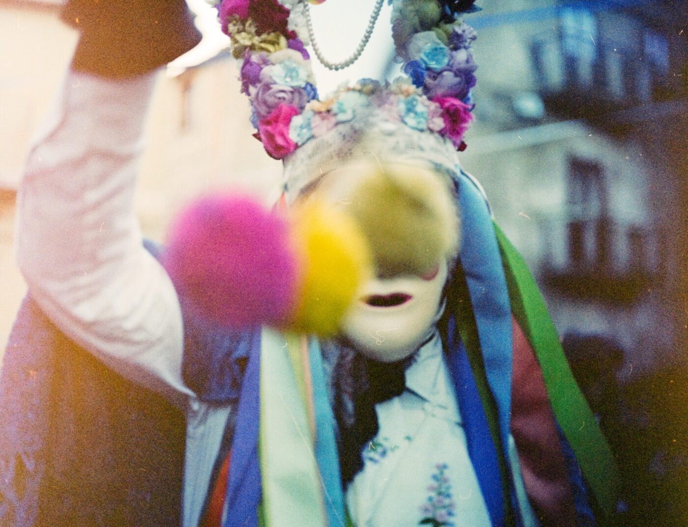 Una persona con una maschera bianca e un costume vivace decorato con fiori, nastri e perline tiene all'esterno dei pom-pom rosa e gialli sfocati.