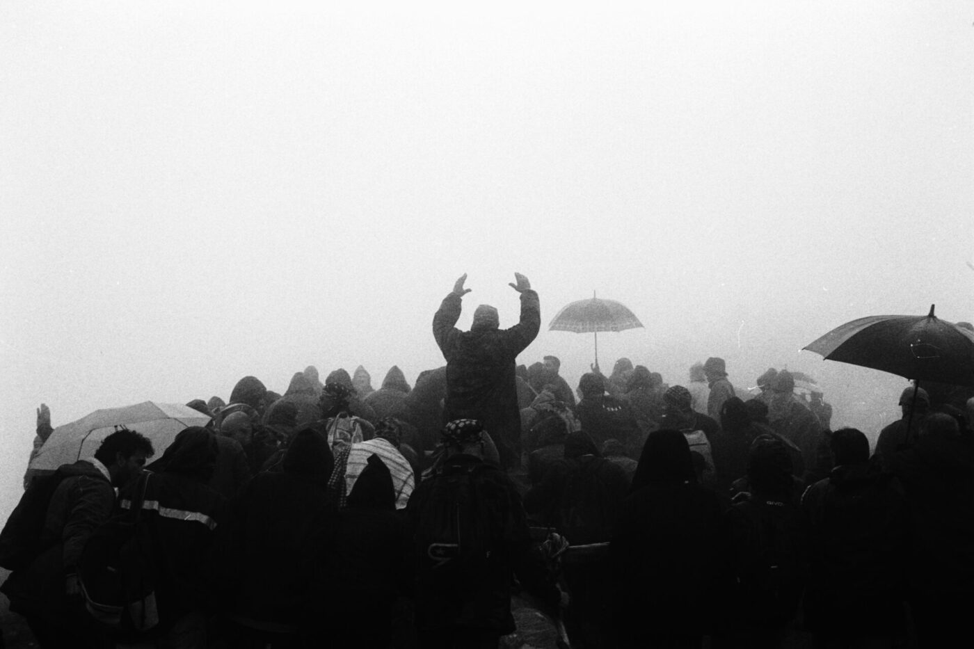 Una folla si raduna nella nebbia fitta, molti con ombrelli, una figura centrale che alza le mani. L'atmosfera è cupa e la visibilità è scarsa.
