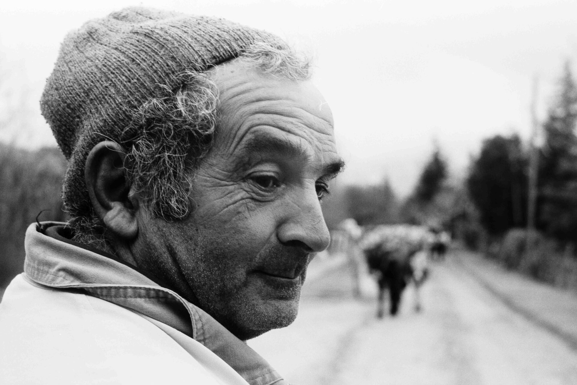Foto in bianco e nero di un uomo anziano con capelli ricci e berretto, che sorride debolmente a sinistra; strada rurale e figure sfocate sullo sfondo.