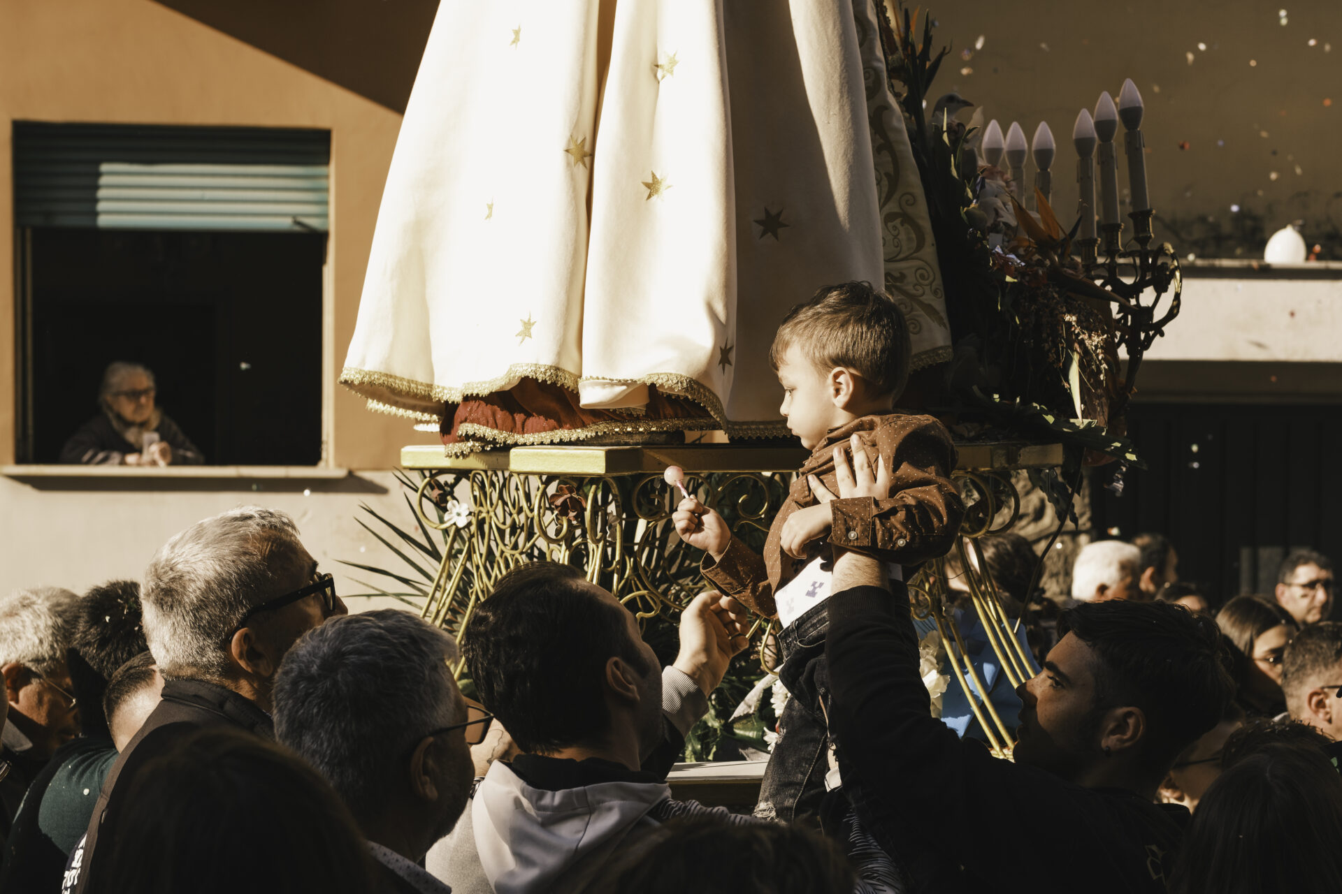 Un gruppo solleva un bambino verso un carro decorato durante un affollato evento all'aperto; la calda luce del sole lo illumina mentre gli spettatori guardano e partecipano.