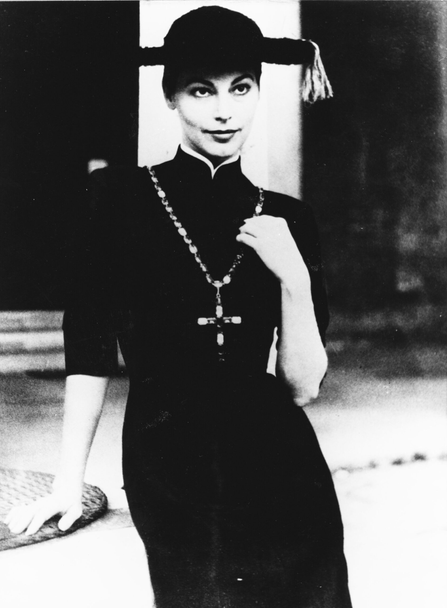 Una donna in abito scuro e cappello con nappe sta in piedi con sicurezza, indossando una vistosa collana di croci. Immagine in bianco e nero, in stile vintage.