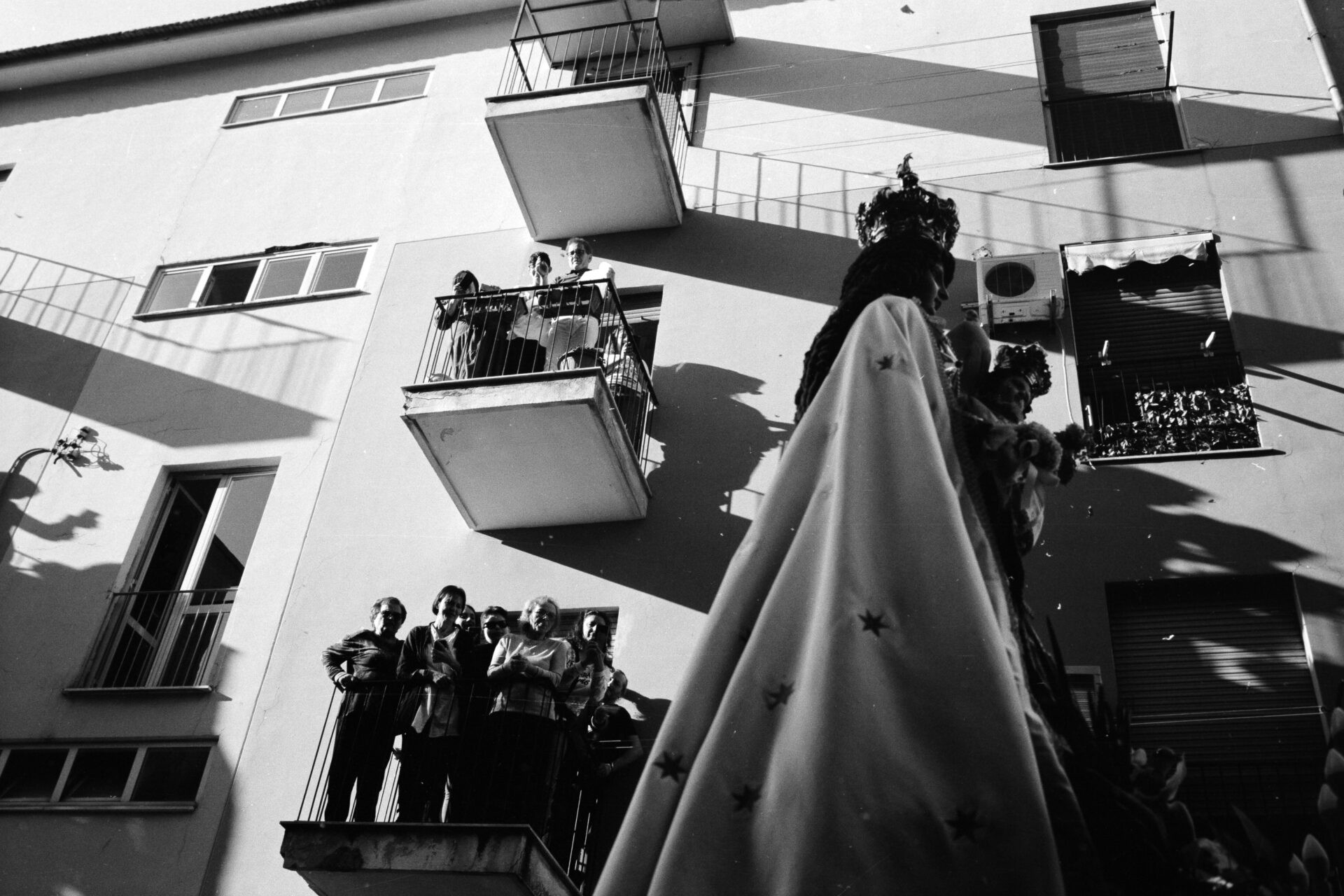 Una statua religiosa con un drappo decorato viene portata davanti agli spettatori sui balconi durante una processione cittadina in bianco e nero.